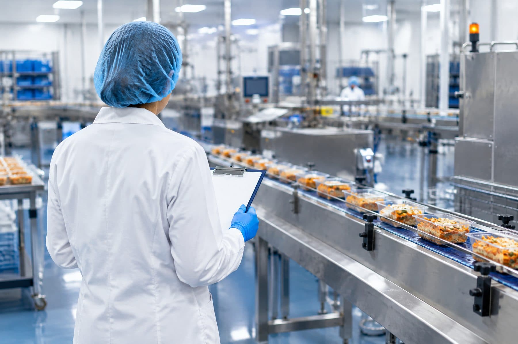 A food technologist in a white coat and hairnet inspecting products on a production line, seen from behind