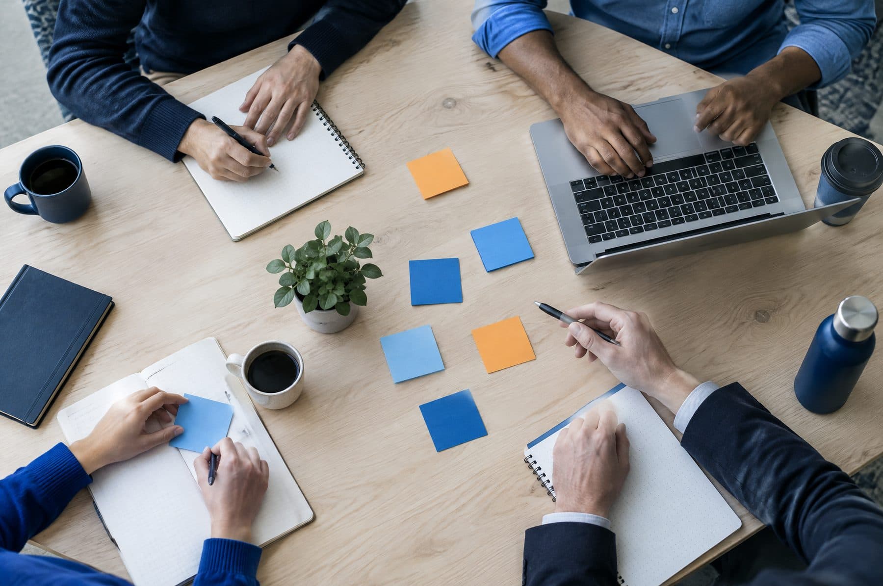 Colleagues' hands around a table with notebooks, a laptop and sticky notes during a planning session